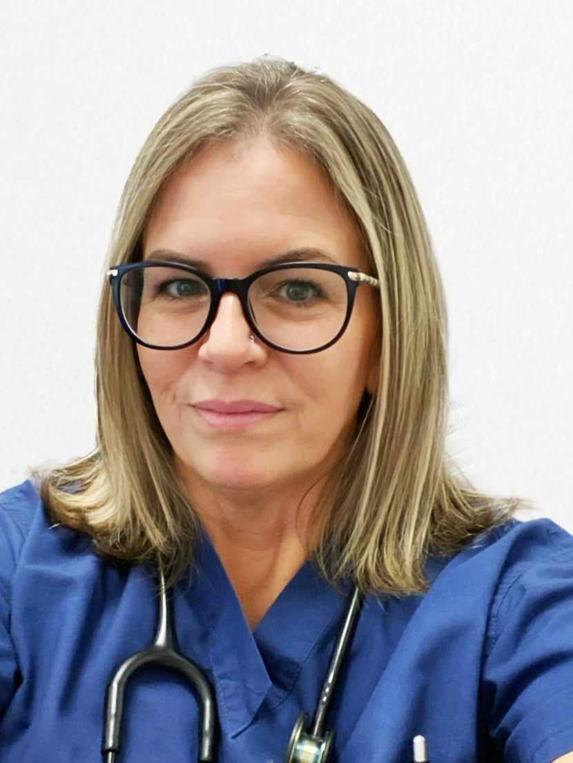 Nicole wearing dark blue scrubs, glasses, and a stethoscope, smiling at the camera.